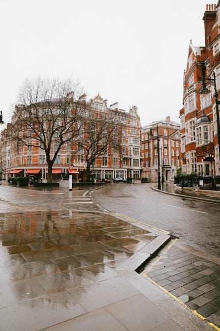 The image depicts a narrow street in Mayfair, showing part of a cream-colored, multi-story building with several windows, some with reflective glass panes. A black outdoor lamp is mounted on the build