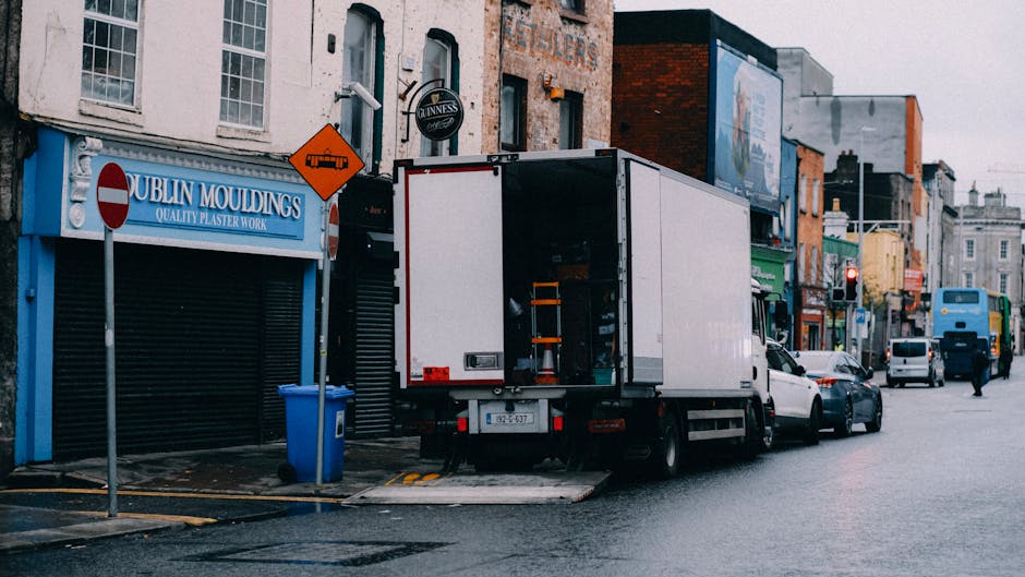 A large green and beige delivery truck is parked on an urban street in front of tall modern glass skyscrapers and older stone buildings. The truck's rear doors are closed, and it is positioned close t