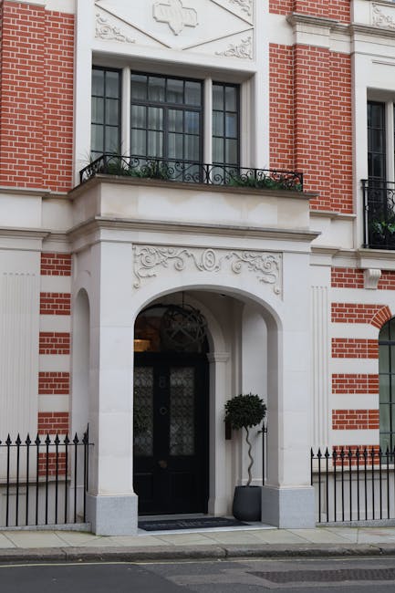 Two movers from Storage Mayfair are preparing to pack and load a white fabric sofa in a well-lit living room with large windows and a potted plant in the background. The woman, dressed in a light blue