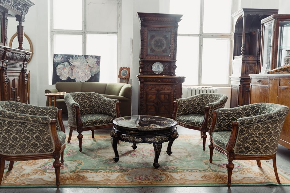 A collection of elegant, antique-style dining chairs with ornate, decorative backrests featuring classical urn motifs are arranged closely together inside a room, possibly a storage area or staging sp