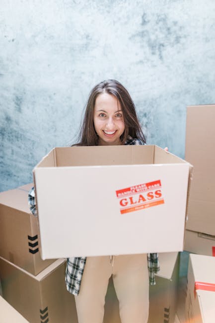 A young woman with long brown hair and a smile is holding a large empty cardboard box labeled 'Glass' with red printing, standing indoors against a textured light blue wall. She is wearing a plaid shi